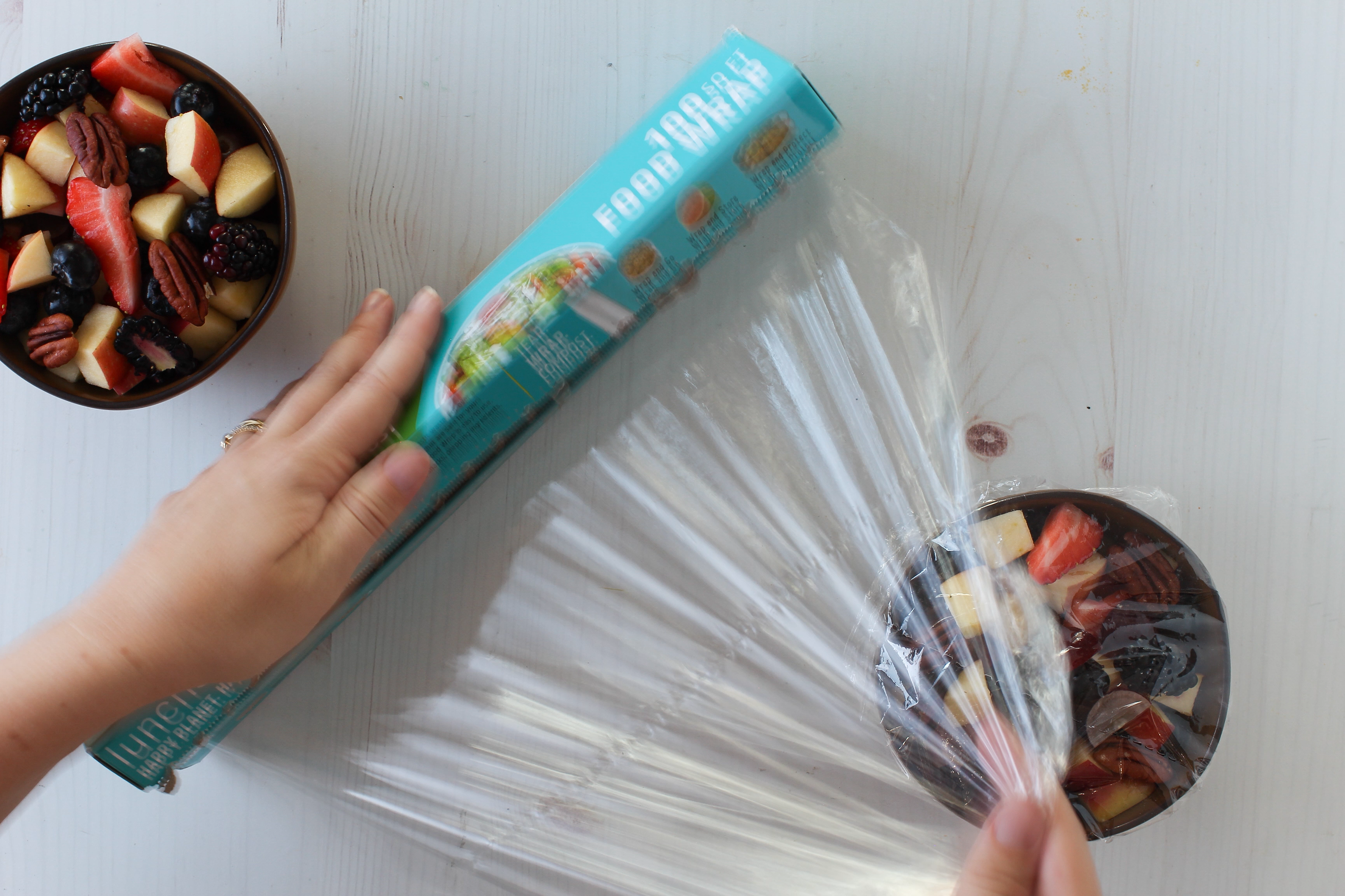 Image of Hand holding a box of fruit snacks next to a bowl of mixed fruits on a white surface.