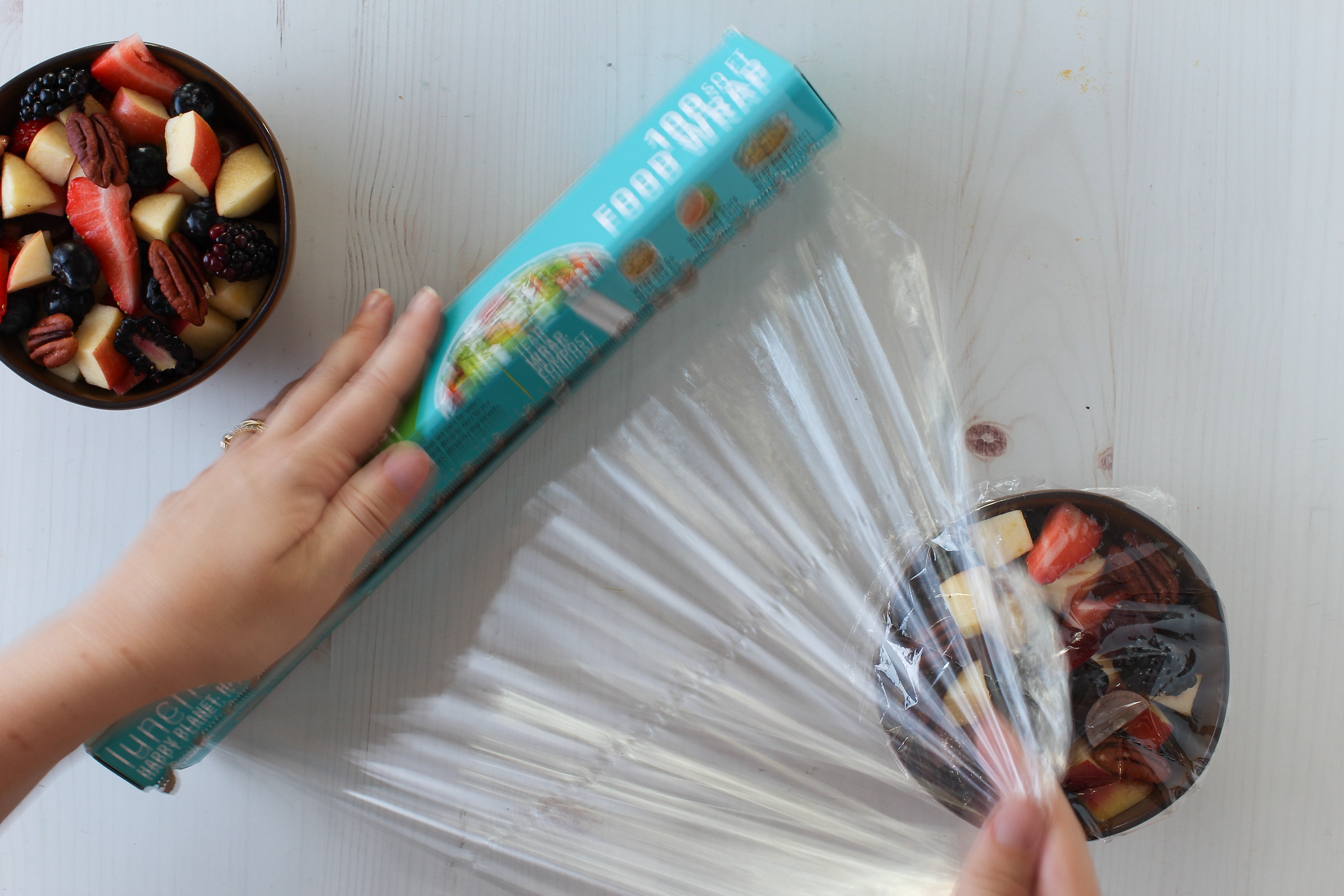 Hand holding a box of fruit snacks next to a bowl of mixed fruits on a white surface.