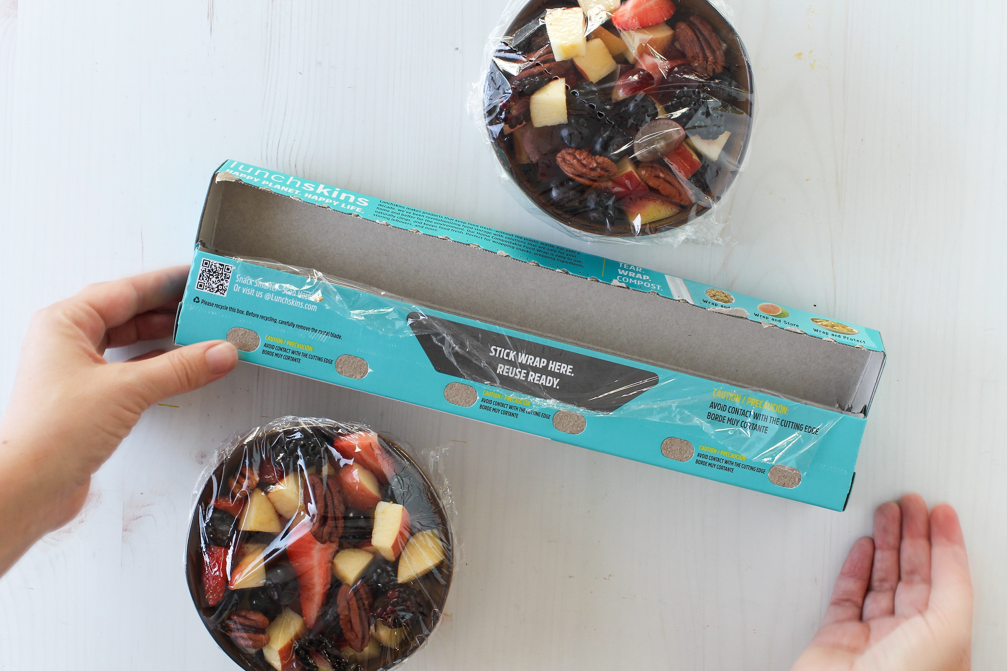 Image of Hands opening a box containing a fruit  with a visible brand logo. Showing Reuse Ready Tab Lunchskins Food Wrap