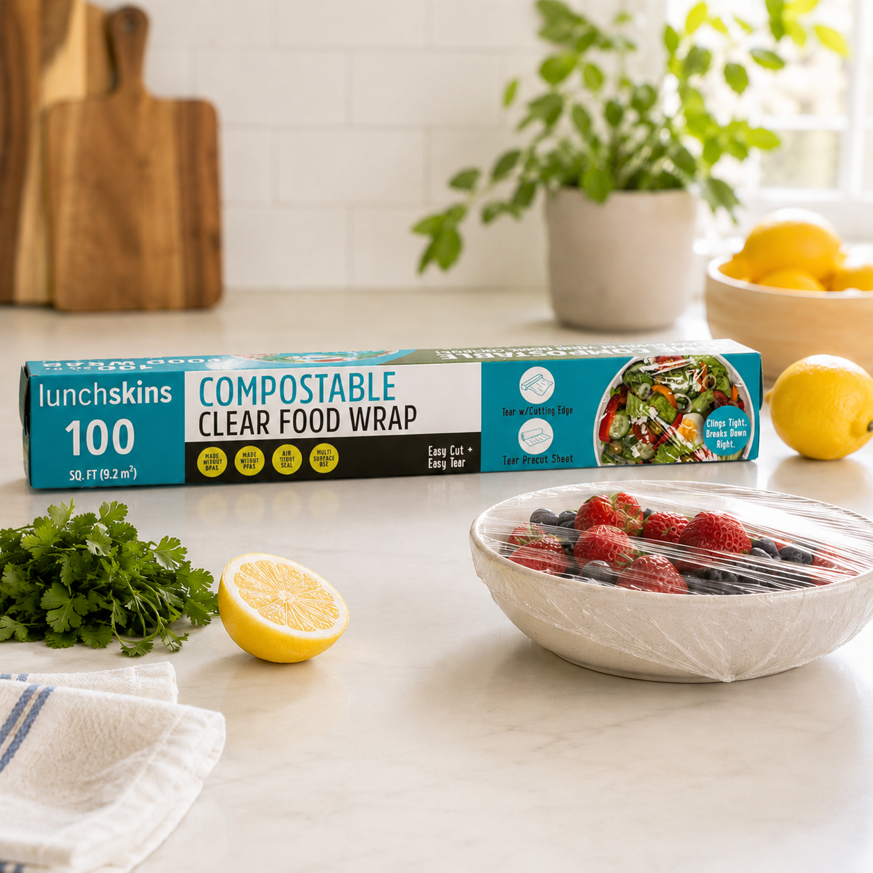Image of Lunchskins compostable clear food wrap box displayed on a bright kitchen counter beside a bowl of fresh berries covered with transparent compostable food wrap, lemons, and fresh herbs.